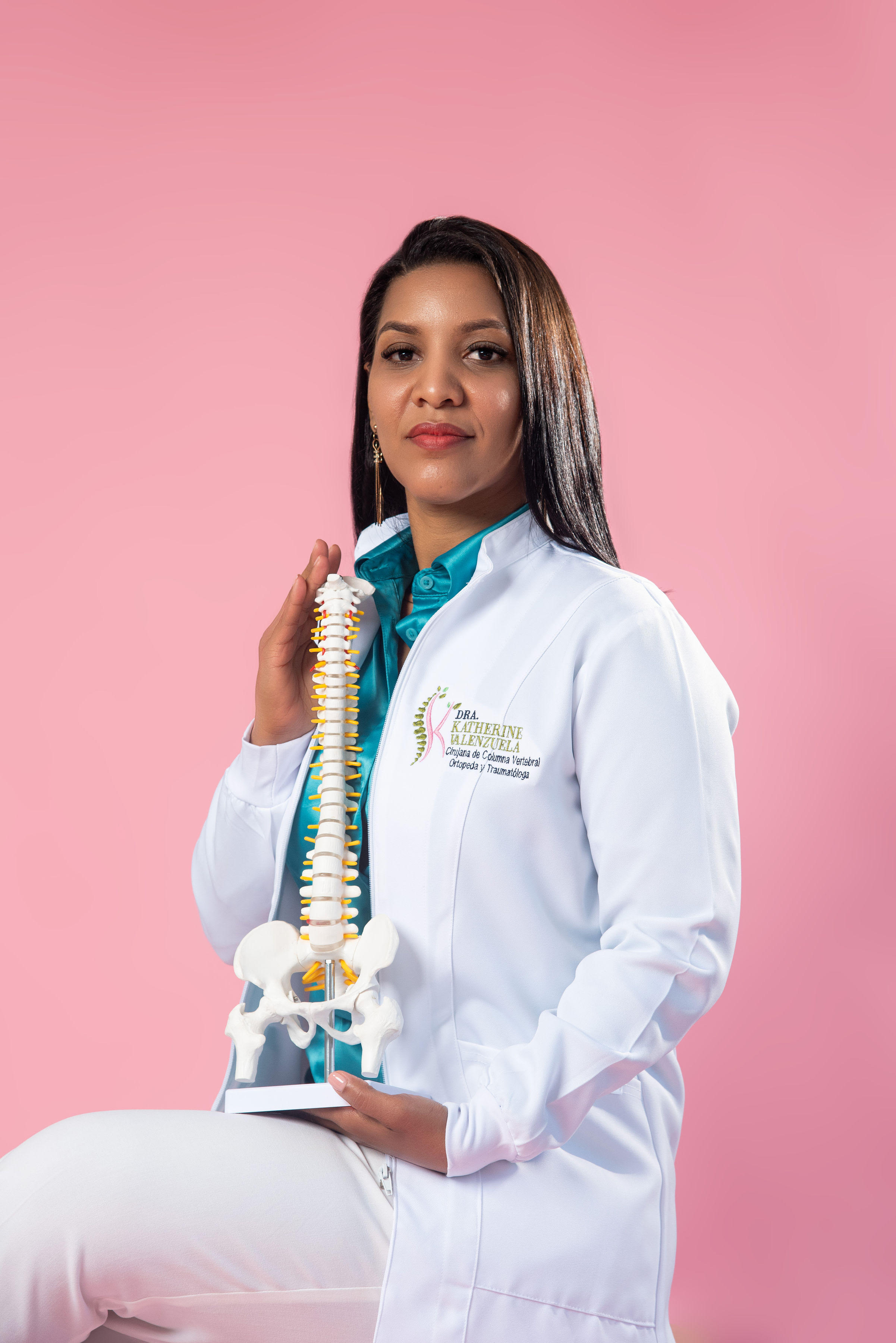 Retrato profesional de la Dra. Katherine Valenzuela con bata blanca sosteniendo un modelo anatómico de columna vertebral frente a un fondo rosa liso, mirando a cámara con expresión serena y confiada.